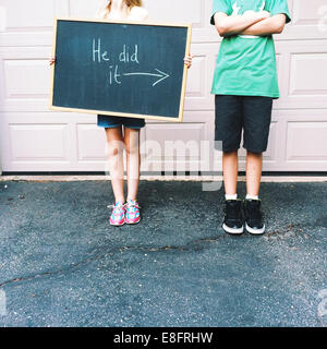 Boy standing next to a girl holding a blackboard saying he did it Stock Photo