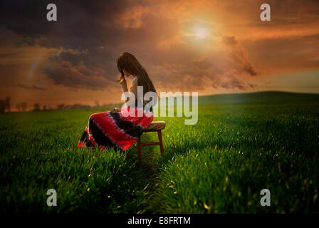 Side view of girl sitting on stool in a meadow at sunset, Poland Stock Photo