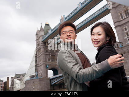 chinese asian couple visiting London (from Singapore) holding each other smiling in front of Tower Bridge Stock Photo