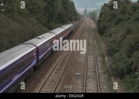 Speeding train carriages run towards signal lights, flanked by green trees and extra sets of tracks. A town in the distance. Stock Photo
