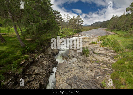 Water of River Dee rushing down through narrow rocky gorge surrounded by emerald green grass and pine forest under blue sky at Linn o' Dee in Scotland Stock Photo