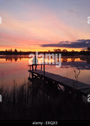 Silhouette of man sitting on jetty by lake at sunset, Gotaland, Sweden Stock Photo
