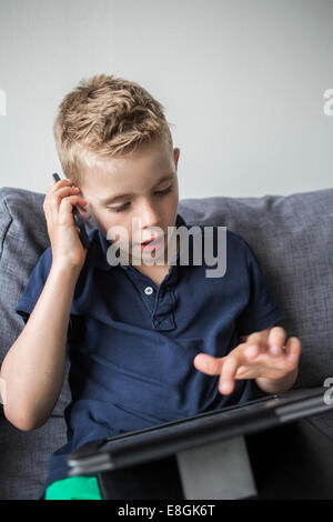 Boy answering smart phone while using digital tablet on sofa at home Stock Photo