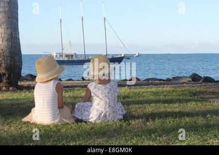 Two girls sitting on grass looking at boat, Port Douglas, Queensland, Australia Stock Photo