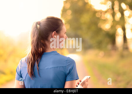 Attractive happy woman standing listening to music on a rural road in sunlight with her MP3 player and earplugs
