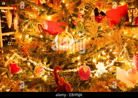 Christmas Tree With Ornaments United States / UNITED STATES — A close-up view of a brightly lit Christmas tree adorned with numerous festive ornaments and warm lights. Golden tinsel and a variety of unique decorations, including a flamingo, a red fish, and a star-patterned bauble, create a vibrant and cozy holiday atmosphere. The tree is also decorated with a red stocking, a red monkey, and a red card. Stock Photo