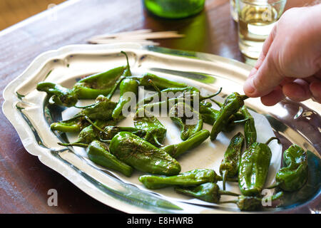 padron peppers as a typical Spanish tapas style snack on wood table , with sherry glasses and bottle in background and hand hold Stock Photo