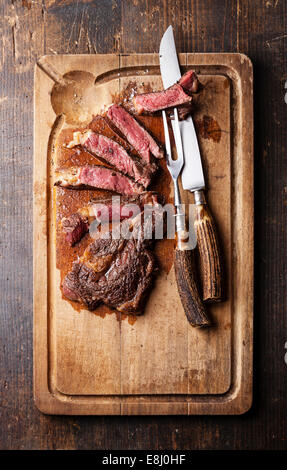 Medium rare Beef steak Ribeye with knife and fork for meat on cutting board on dark wooden background Stock Photo