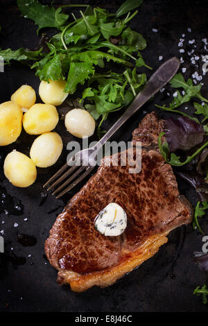Grilled steak with butter, potatoes and green salad over black metal board Stock Photo