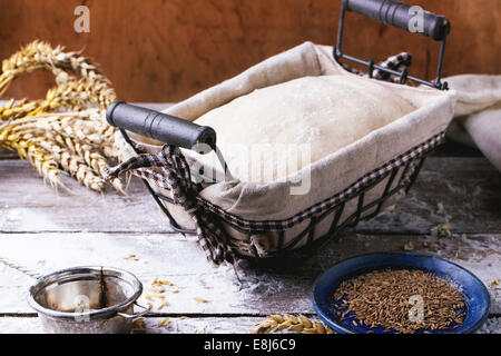 Baking bread. Dough in proofing basket on wooden table with flour, cumin and wheat ears. See series Stock Photo