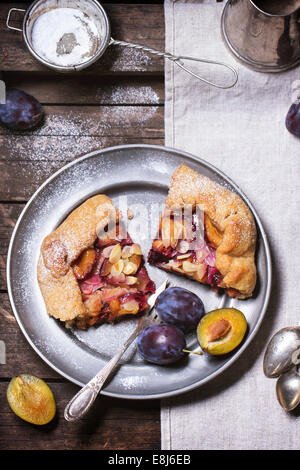 Galette cake with plums, served in vintage metal plate over old wooden table. Stock Photo