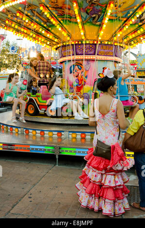 Spanish woman in traditional festive dress waiting next to chlidrens Carousel, merry-go-round at annual fair. Fuengirola, Spain. Stock Photo