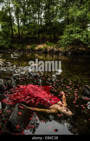 Modern Ophelia: A woman girl wearing deep red frock ball gown lying on her back surrounded by petals floating 'dead' in river