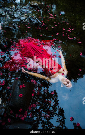 Modern Ophelia: A woman girl wearing deep red frock ball gown lying on her back surrounded by petals floating 'dead' in river
