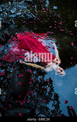 Modern Ophelia: A  woman girl wearing deep red frock ball gown lying on her back surrounded by petals floating 'dead' in river Stock Photo
