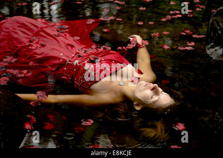 Modern Ophelia: A  woman girl wearing deep red frock ball gown lying on her back surrounded by petals floating 'dead' in river Stock Photo