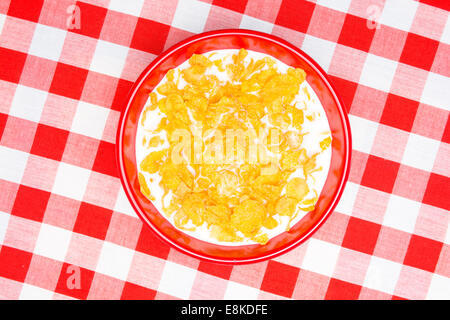 A red bowl of cornflakes and milk on a classic, red, checkered tablecloth ready as a breakfast meal. Stock Photo