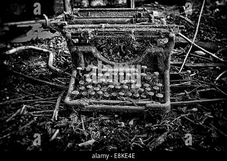An abandoned and broken, antique, vintage typewriter, left outdoors with weeds and grass growing over it. In black and white. Stock Photo