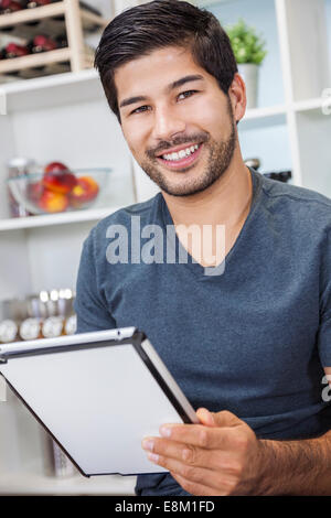 Handsome smiling young Asian man with a beard using tablet computer at home in his kitchen Stock Photo