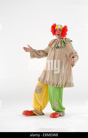 man dressed in fancy dress comedy costume in a circus clown outfit with funny wig, baggy clothes, giant shoes & red nose Stock Photo