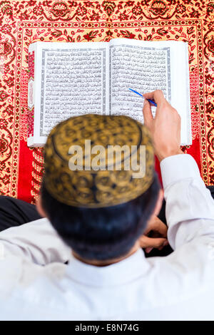 Asian Muslim man reading Koran or Quran on praying carpet wearing traditional dress Stock Photo