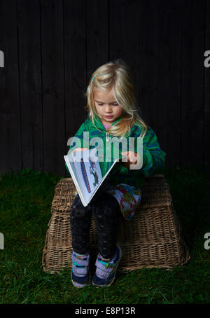 little child blond girl sitting on a wicker basket is reading a book outdoors, dark wooden wall background Stock Photo