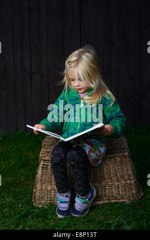 little child blond girl sitting on a wicker basket is reading a book outdoors, dark wooden wall background Stock Photo
