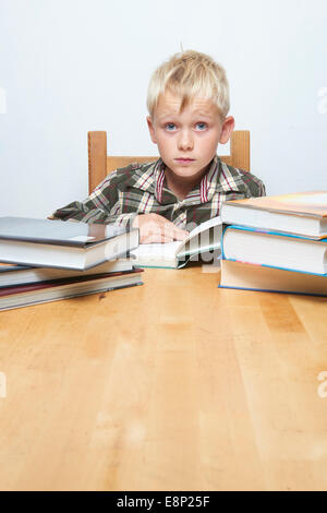 Little child blond focused student boy sitting at desk and reading book, more books at table around,  white background Stock Photo