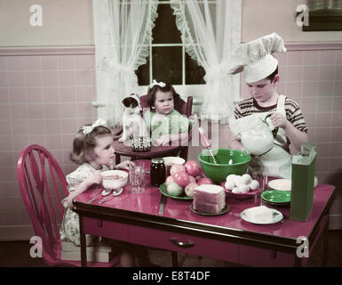 1940s 1950s BOY TWO GIRLS AT KITCHEN TABLE BOY IN TOQUE COOKING ONE GIRL EATING TODDLER AND PUPPY IN HIGH CHAIR Stock Photo
