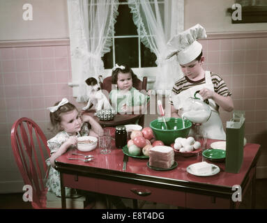 1940s 1950s CHILDREN TWO GIRLS AT KITCHEN TABLE WITH PUPPY AND BOY COOKING WEARING CHEFS TOQUE Stock Photo