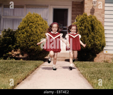 1960s TWIN GIRLS WEARING IDENTICAL RED DRESSES HOLDING HANDS RUNNING DOWN SIDEWALK OF SUBURBAN HOUSE LOOKING AT CAMERA Stock Photo
