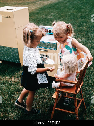 1960s TWO LITTLE GIRLS PLAYING TOGETHER FEEDING DOLL TOY STOVE REFRIGERATOR IN BACKYARD Stock Photo