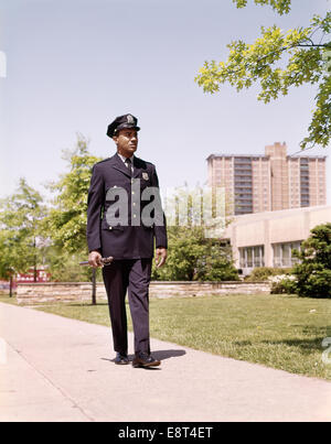 1960s AFRICAN AMERICAN POLICEMAN OFFICER WALKING SIDEWALK NEIGHBORHOOD BEAT Stock Photo