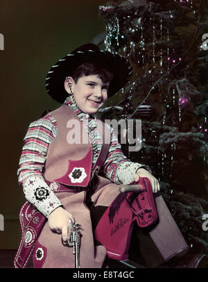 1940s 1950s SMILING BOY DRESSED IN COWBYO HAT COSTUME HOLDING TOY GUN SITTING WOODEN HORSE BY CHRISTMAS TREE Stock Photo