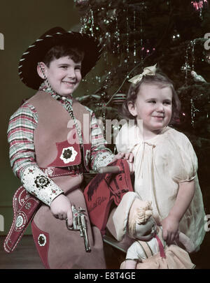 1950s SMILING BOY DRESSED IN COWBOY HAT OUTFIT HOLDING CAP PISTOL LITTLE GIRL HOLDING A DOLL BY CHRISTMAS TREE Stock Photo