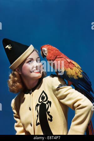 1940s SMILING BLOND WOMAN WEARING YELLOW AND BLACK JACKET AND POINTED HAT LOOKING AT PARROT ON SHOULDER Stock Photo