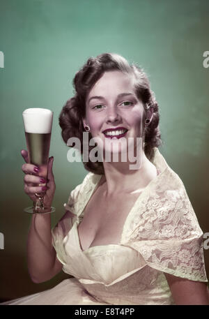 1950s PORTRAIT SMILING WOMAN WEARING FORMAL GOWN HOLDING PILSNER GLASS OF BEER LOOKING AT CAMERA Stock Photo