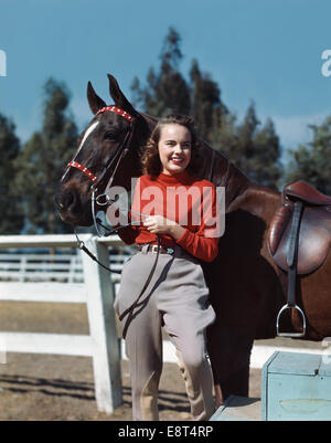 1940s 1950s SMILING TEEN GIRL WEARING JODHPURS POSING WITH HORSE BY FENCE Stock Photo