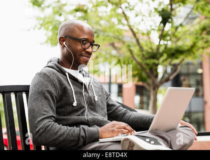 Black man using laptop and earphones on city bench Stock Photo