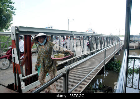 a local Vietnamese lady in traditional hat walks across a bridge on a river crossing carrying her goods Stock Photo