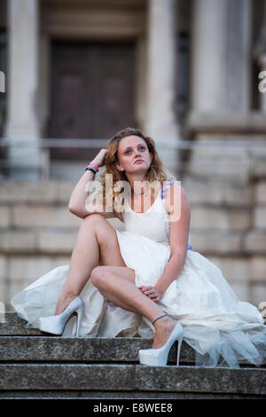 The runaway or jilted bride - a young woman girl in a wedding dress sitting alone sad on stone stairway, evening outdoors uk