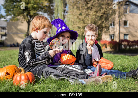 Three little cute friends sitting on the grass and eating Halloween candies Stock Photo