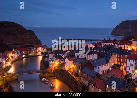 Evening view of quaint seaside cottages (lights on) towering cliffs & harbour of old fishing village - Staithes, North Yorkshire Coast, England, UK. Stock Photo