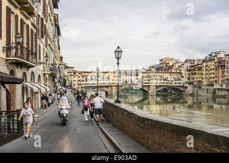 pedestrians crowd the Lungarno Degli Acciaiuoli on North bank of the Arno overlooking the Ponte Vecchio & calm river waters Stock Photo