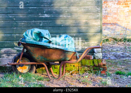 Old rusty wheelbarrow standing in a garden next to a green shed waiting to be used.