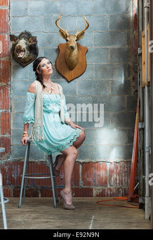 Young Woman in Bohemian Clothes Sitting on Stool Next to Two Animal Heads Hanging on Wall, Portrait Stock Photo