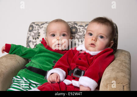 fraternal twin 9 month old baby boys wearing a santa and an elf suit on their first Christmas, both sitting on a chair Stock Photo