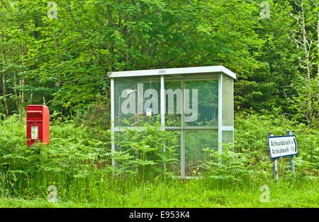 Post box and shabby little-used bus shelter surrounded by tall bracken and grass at road junction in woodland near Plockton Stock Photo