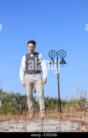 man in shirt and vest with bow tie and glasses, standing in lines of railway in the old vintage park and looking at camera Stock Photo