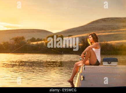 Young woman fishing from a dock on a small lake at sunset. Stock Photo